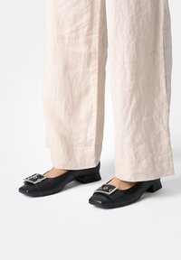 Chaussures noires en cuir à talons blocs avec un bout arrondi et un large détail boucle argentée sur le devant, associées à un pantalon large de couleur claire.