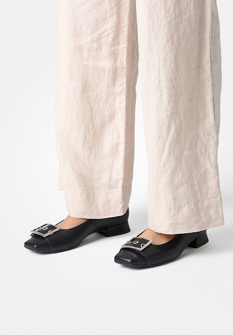 Chaussures noires en cuir à talons blocs avec un bout arrondi et un large détail boucle argentée sur le devant, associées à un pantalon large de couleur claire.