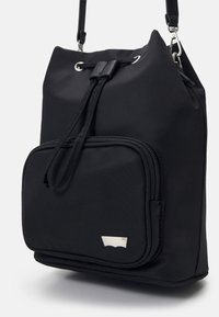 Czarny nylonowy plecak z ściąganym zamknięciem, przednia kieszeń zapinana na zamek oraz małe srebrne logo w dolnym prawym rogu przedniej kieszeni.