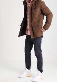 Chaqueta de algodón marrón con botones, bolsillos laterales, llevada sobre una sudadera rosa. Jeans oscuros y zapatillas blancas con acentos beige completan el conjunto.