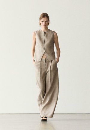 Woman walking indoors wearing beige sleeveless buttoned vest and wide-leg trousers with hands in pockets and black sandals.