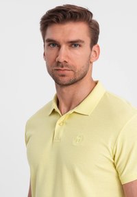 Lysgul polo T-shirt med krave, tre knapper og et struktureret stof. Har et prægede logo på brystet.