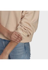 Beige sweatshirt med mjuk textur och avslappnad passform; har rullade ärmar och jeans i denim med en urtvättad look. Händerna vilar på armen.