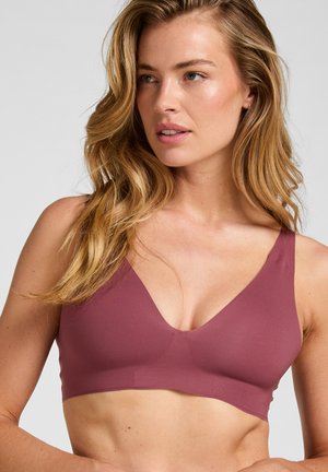 Vrouw met lang golvend blond haar die een naadloze mauve bh-top draagt, iets naar links kijkend tegen een effen lichte achtergrond.