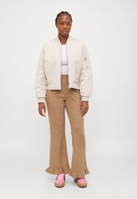 Light beige bomber jacket, white top, tan ribbed flared pants with ruffle hem, and pink sneakers. Smooth fabric and relaxed fit.