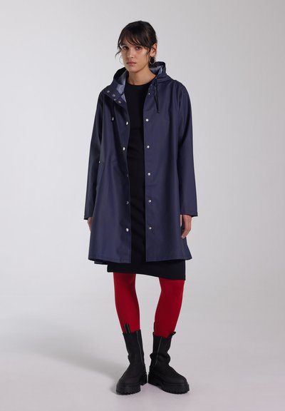 Navy waterproof raincoat with a hood, snap closures, and side pockets, paired with a black dress and red tights, featuring chunky black boots.