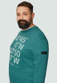 Sweatshirt bleu canard, col ras du cou, avec des coordonnées géographiques blanches imprimées sur le devant et une étiquette tissée noire sur la manche.