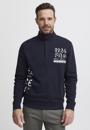 Marineblå genser med kvart glidelås, broderte grafikker i hvitt og rødt, ribbede mansjetter, myk tekstur og avslappet passform.