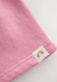 Esquina de tela rosa suave con dobladillo cosido y una pequeña etiqueta beige con un icono minimalista de arcoíris en tonos neutros.