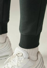 Dunkelgrüne Jogginghosen mit gerippten Bündchen, kombiniert mit weißen Sportschuhen und strukturierten Crew-Socken. Fokussiert auf Form- und Materialdetails.