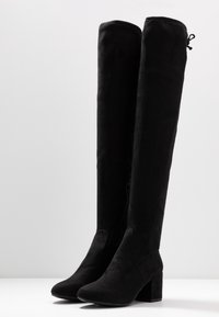 Botas negras por encima de la rodilla hechas de suave gamuza, con forma cilíndrica, tacones cuadrados, cierre de cremallera y un detalle de encaje en la parte superior.