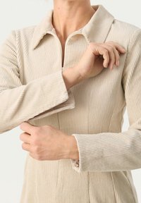 Veste en velours côtelé beige avec fermeture éclair à l'avant, dotée d'un col avec revers et d'une texture côtelée. Les manches ont une coupe décontractée, mettant en valeur des coutures discrètes.