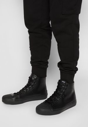 Czarne wysokie trampki na sznurowadła noszone z czarnymi spodniami dresowymi na jasnym, jednolitym tle.