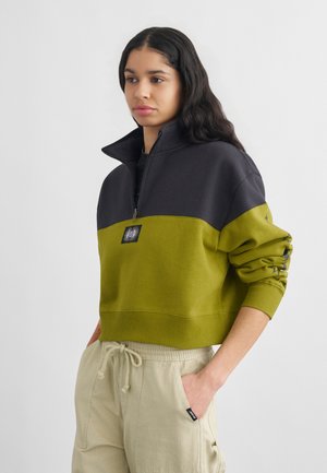 Mladá žena s dlhými tmavými vlasmi, oblečená v čiernej a olivovo zelenej mikine so štvrťzipsom a béžových nohaviciach so šnúrkou, ruky v bodoch.