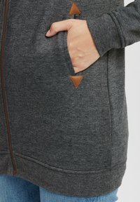 Kolgrå zip-up sweatshirt med texturerad tyg, sidofickor och bruna triangulära läderdetaljer nära fickorna.