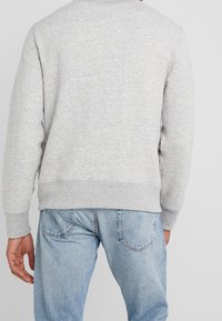 Grå sweatshirt med rund hals, ribbade manschetter och nederkant. Mjuk textur, enkel design, bärs med ljusblå jeans. Synliga sömnadsdetaljer.