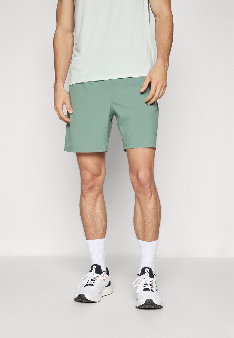 Menns atletiske antrekk med lysegrå skjorte, grønne shorts og hvite sokker med svarte og grå joggesko. Shortsene har en glatt tekstur.