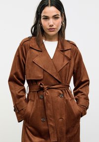 Hnědý semišový trench coat s páskem v pase, velkými klopy, dvouřadým zapínáním a bočními kapsami. Hladká textura s uvolněným střihem.