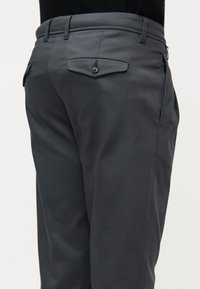 Pantalon gris foncé sur mesure avec une poche arrière à rabat boutonnée, des poches latérales, des passants de ceinture, et un haut en maille noire rentré à l'intérieur.