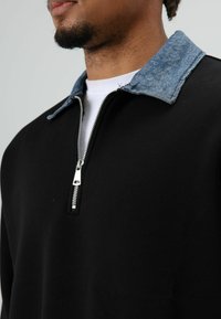 Zwarte sweatshirt met een ritssluiting bij de kraag, voorzien van een contrasterend blauw denim accent. Gladde textuur met glanzende zilveren ritssluitingdetail.
