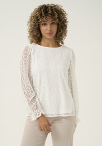 White long-sleeve blouse featuring a textured pattern with diamond shapes, sheer lace accents on sleeves, and a crew neckline.
