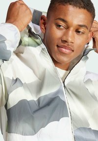 Lightweight jacket in a gray and green wave pattern. Features a high collar, zip closure, and attached hood. Smooth, water-resistant fabric.