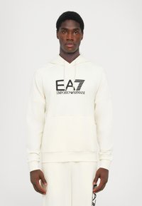 Felpa crema realizzata in tessuto morbido, con tasca a marsupio davanti, polsini a costine e logo nero "EA7 Emporio Armani".