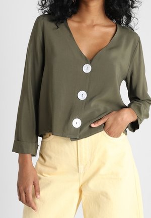 Kvinde iført en olivengrøn V-hals bluse med tre store hvide knapper og beige bukser med høj talje, den ene hånd i lommen.