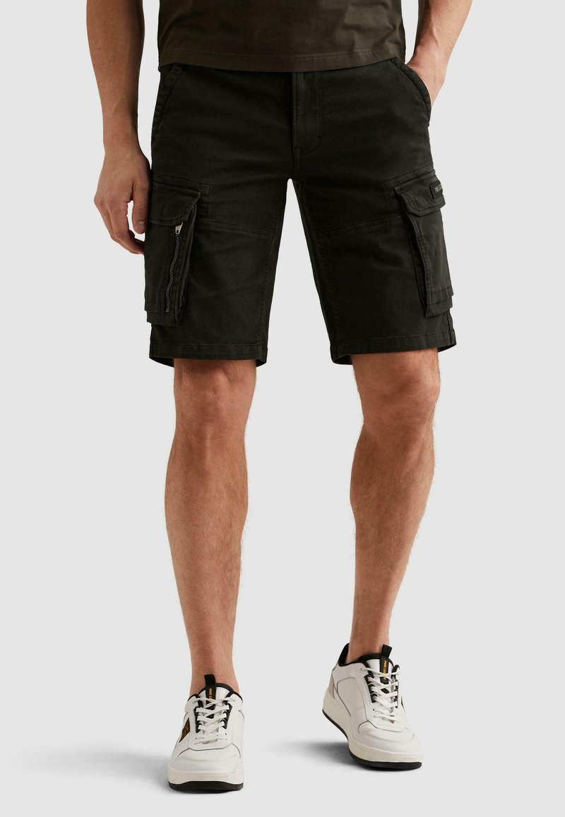 Homme portant un short cargo noir avec rabats et poches zippées, des baskets blanches et une chemise sombre sur un fond clair uni.