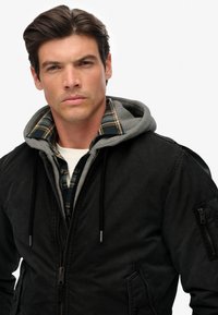 Sort bomberjakke til mænd med en grå hætteklædning under, der har en plaid flannel skjorte. Med lynlås lommer og justerbar snoretræk.