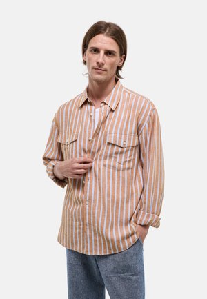 Homme aux cheveux mi-longs portant une chemise beige et blanche à rayures verticales et un pantalon bleu clair, main ajustant le devant de la chemise.
