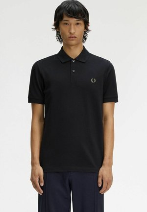 Joven con cabello lacio y negro que lleva una camisa polo negra de manga corta con un pequeño logo bordado en el pecho izquierdo.