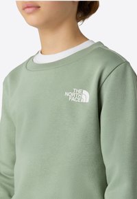 Svetlozelená mikina vyrobená z bavlneného zmesi, so žebrovaným výstrihom a bielym logom na hrudi. Mäkká textúra s voľným strihom.