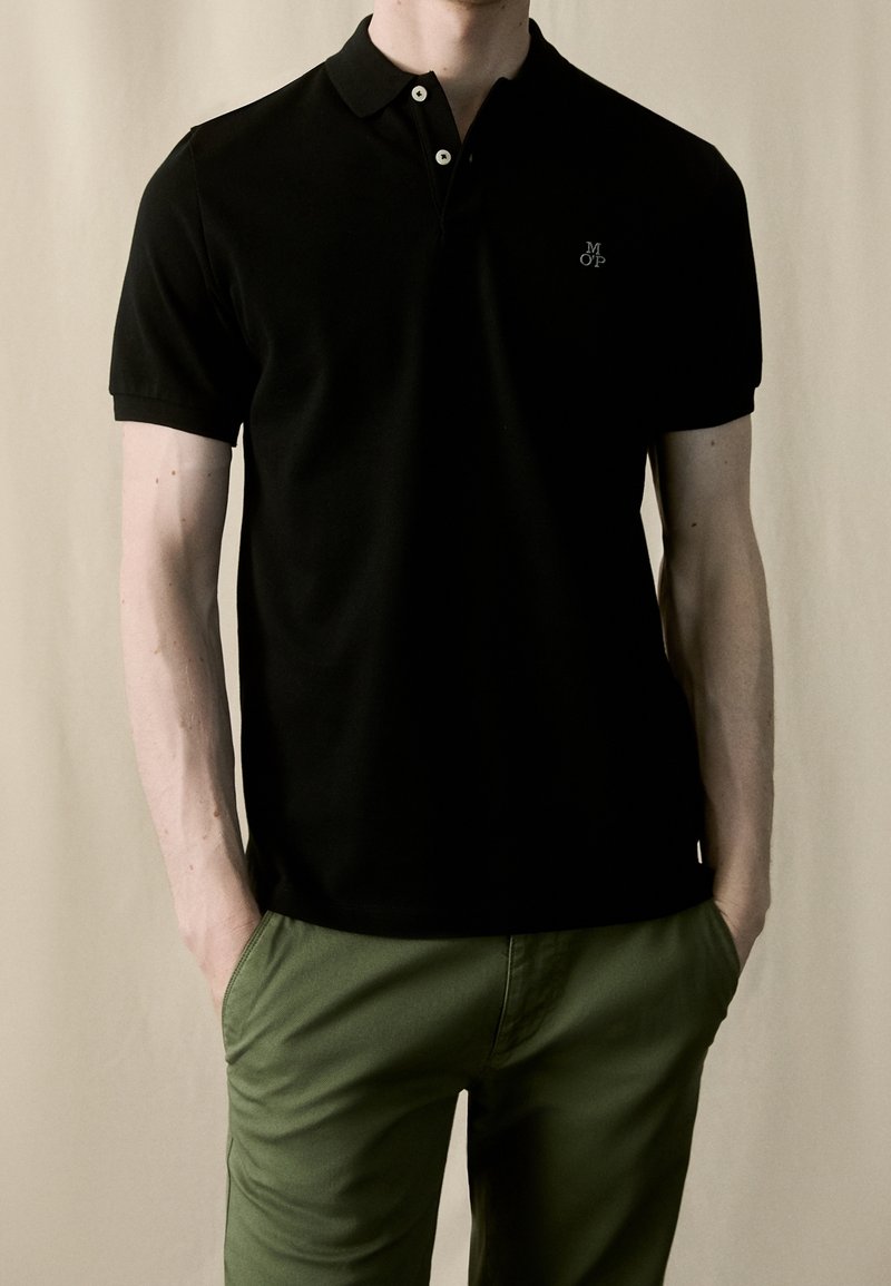 Homme portant un polo noir à manches courtes avec un petit logo blanc sur la poitrine et un pantalon vert olive, les mains dans les poches.