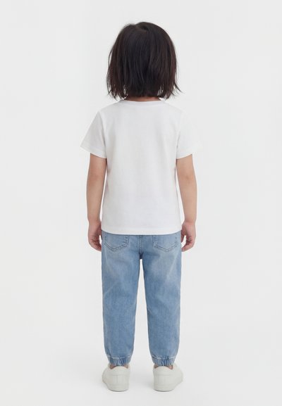 Enfant aux cheveux bruns mi-longs portant un t-shirt blanc uni, un jean bleu clair avec poignets élastiques et des baskets blanches, debout de dos.