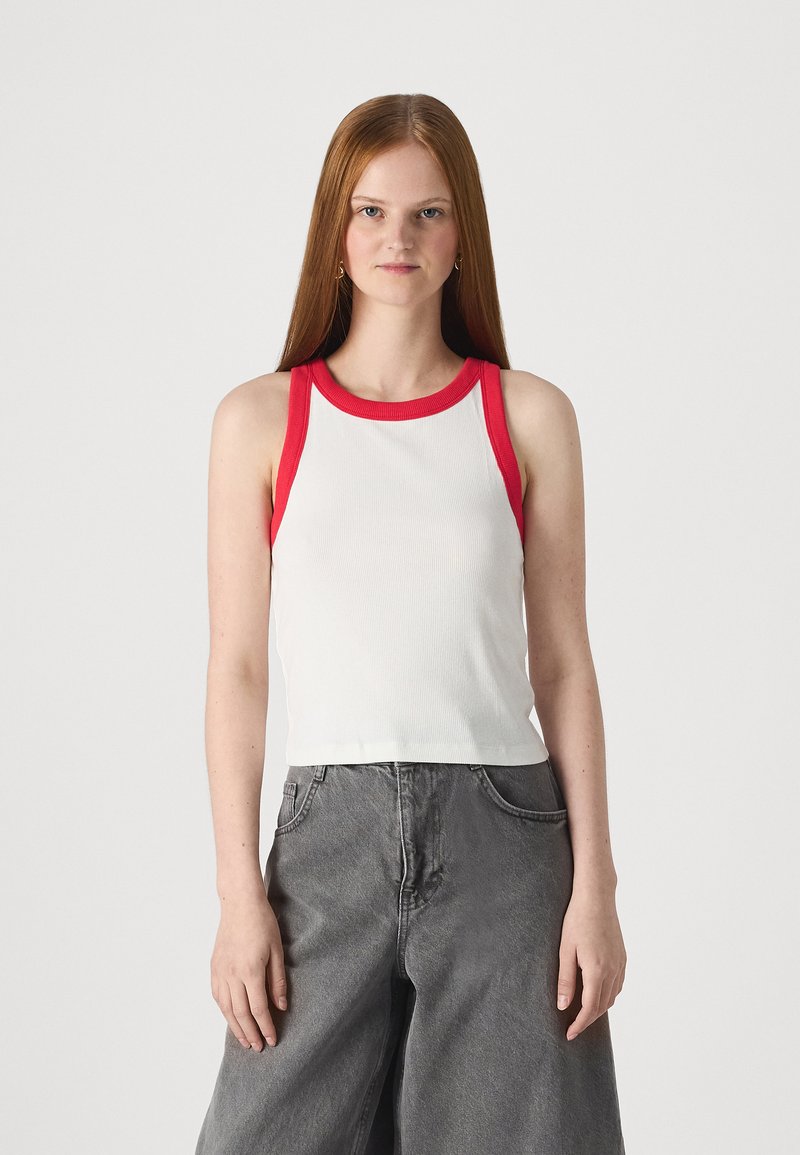 Débardeur blanc à rayures avec des bords rouges, présentant un design ajusté et sans manches. Associe à un jean gris à jambes larges.