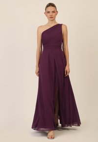 Lila Chiffon-Kleid mit einem Träger, plissiertem Oberteil, fließendem Rock und schulterhohem Schlitz. Sichtbare silberne Riemchen-Absatzschuhe.