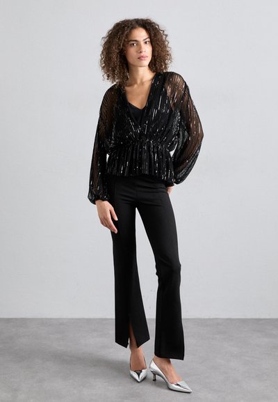 Blouse en sequins noirs avec un décolleté en V et taille cintrée, associée à un pantalon noir avec des fentes latérales, porté avec des talons aiguilles argentés.