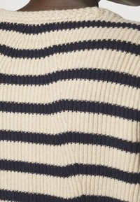 Neulepaita, jossa on tummansinisiä ja krem värisiä vaakaraitoja, ja jossa on teksturoitu ribbikuvio ja pyöreä pääntie.
