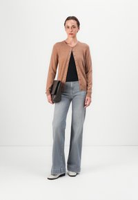 Beige kardigan s knoflíky, černé tričko a světle modré široké džíny. Doplňky zahrnují tmavě hnědou geometrickou kabelku a bílé boty.