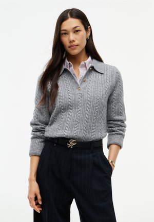Donna che indossa un maglione grigio a trecce sopra una camicia a righe, pantaloni neri a righe sottili e una cintura nera con fibbia dorata, in piedi con una mano in tasca.