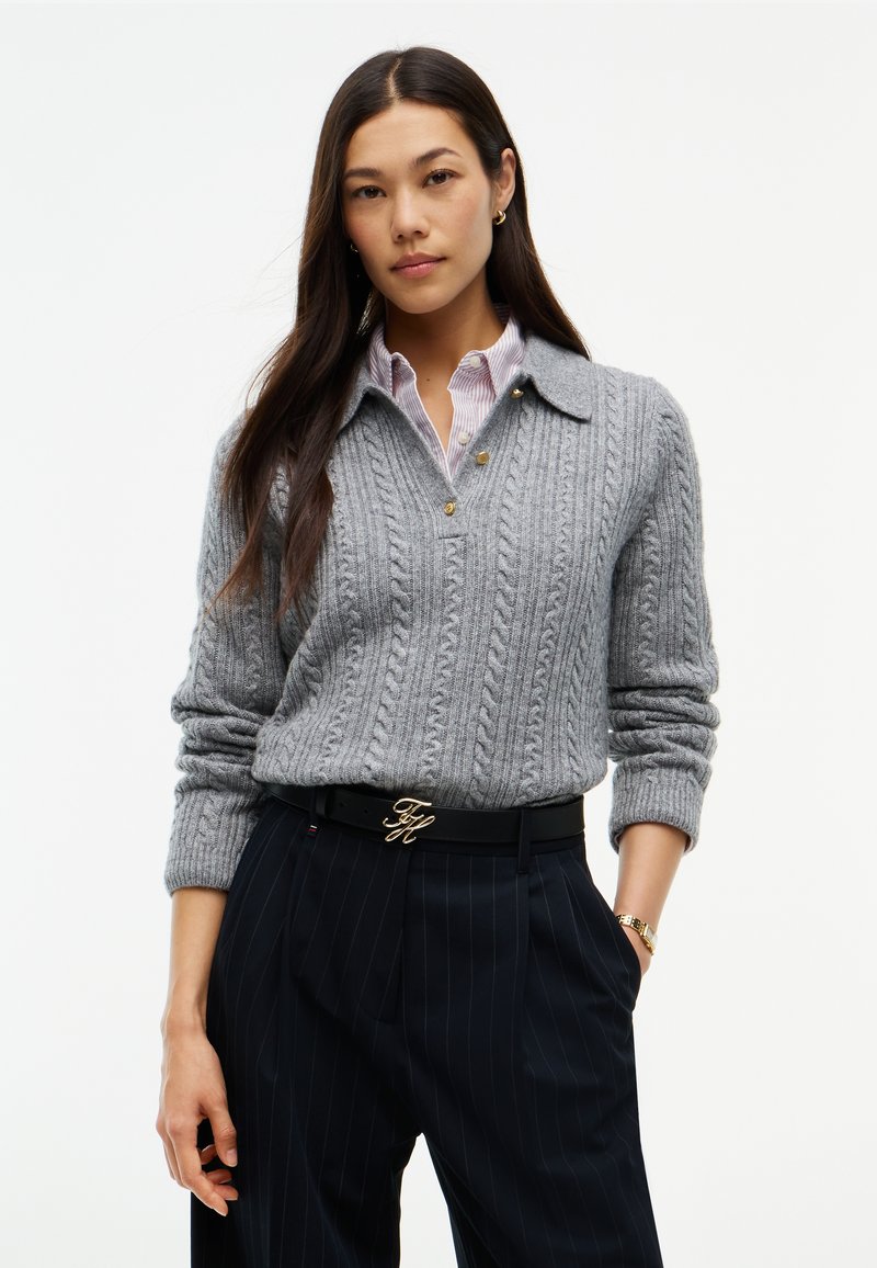 Femme portant un pull gris torsadé par-dessus une chemise à rayures, un pantalon noir à fines rayures et une ceinture noire avec une boucle dorée, debout avec une main dans la poche.