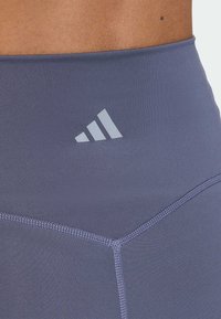 Purple high-waisted athletic leggings with a smooth texture, featuring tonal stitching and a subtle reflective logo on the waistband.