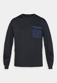 Tricou negru cu mânecă lungă, dintr-un material moale, cu un buzunar albastru pe piept, gât rotund și mâneci cu manșete. Prezintă fermoare cu detalii pe laterale.