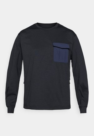 Mustat pitkähihainen t-paita pehmeästä kankaasta, jossa on sininen rintatasku, pyöreä pääntie ja käänteet hihansuissa. Sisältää sivuseinävetoketjut.