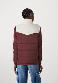 Pufferjack met een bordeauxrode torso en crèmekleurige schouderzone, hoge kraag, gestikte details en ribgebreide manchetten.