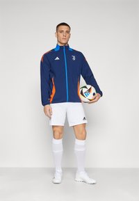 Giacca blu e arancione con zip, pantaloni corti bianchi e calze alte fino al ginocchio. Tiene un pallone da calcio patternizzato. Logo Adidas visibile.