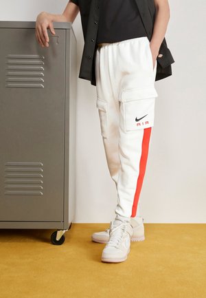 Personne portant un pantalon cargo blanc avec une bande rouge et le logo Nike Air, assorti à des baskets blanches, appuyée sur un casier gris à roulettes.