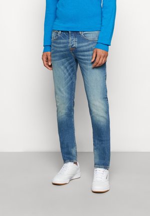 Mann trägt verblasste blaue Jeans, weiße Turnschuhe und ein leuchtend blaues Langarmhemd und steht vor einem einfarbigen weißen Hintergrund.