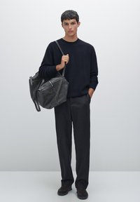 Schwarzer Strickpullover, dunkelgraue Hose, hält eine große schwarze Lederhandtasche mit Reißverschluss und Riemen. Einfaches, strukturiertes Design, matte Oberfläche.